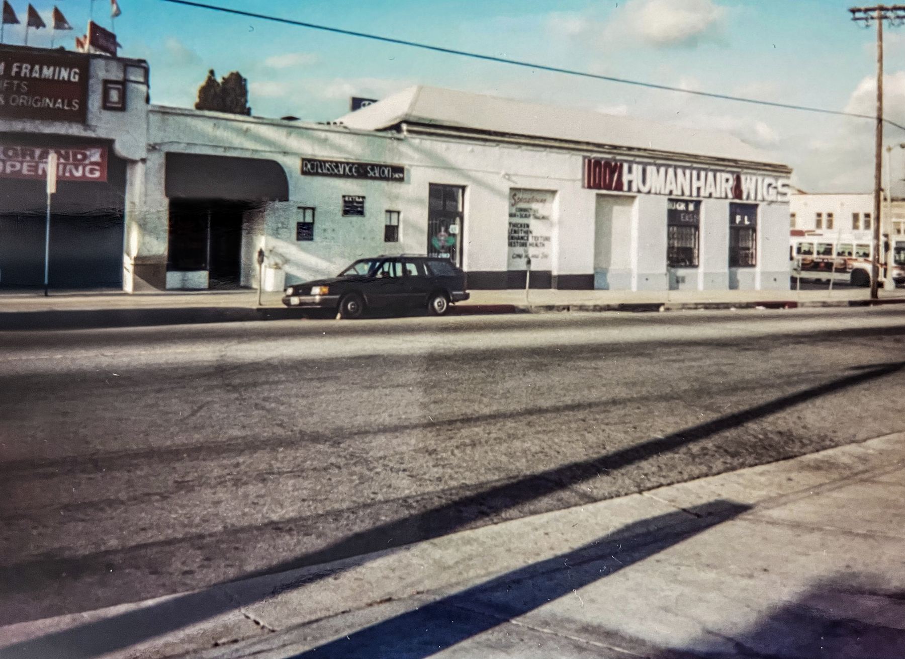 The same building in the 1990s: a single-story white storefront with a Renaissance Salon sign and a 100% Human Hair & Wigs sign, a station wagon parked out front.