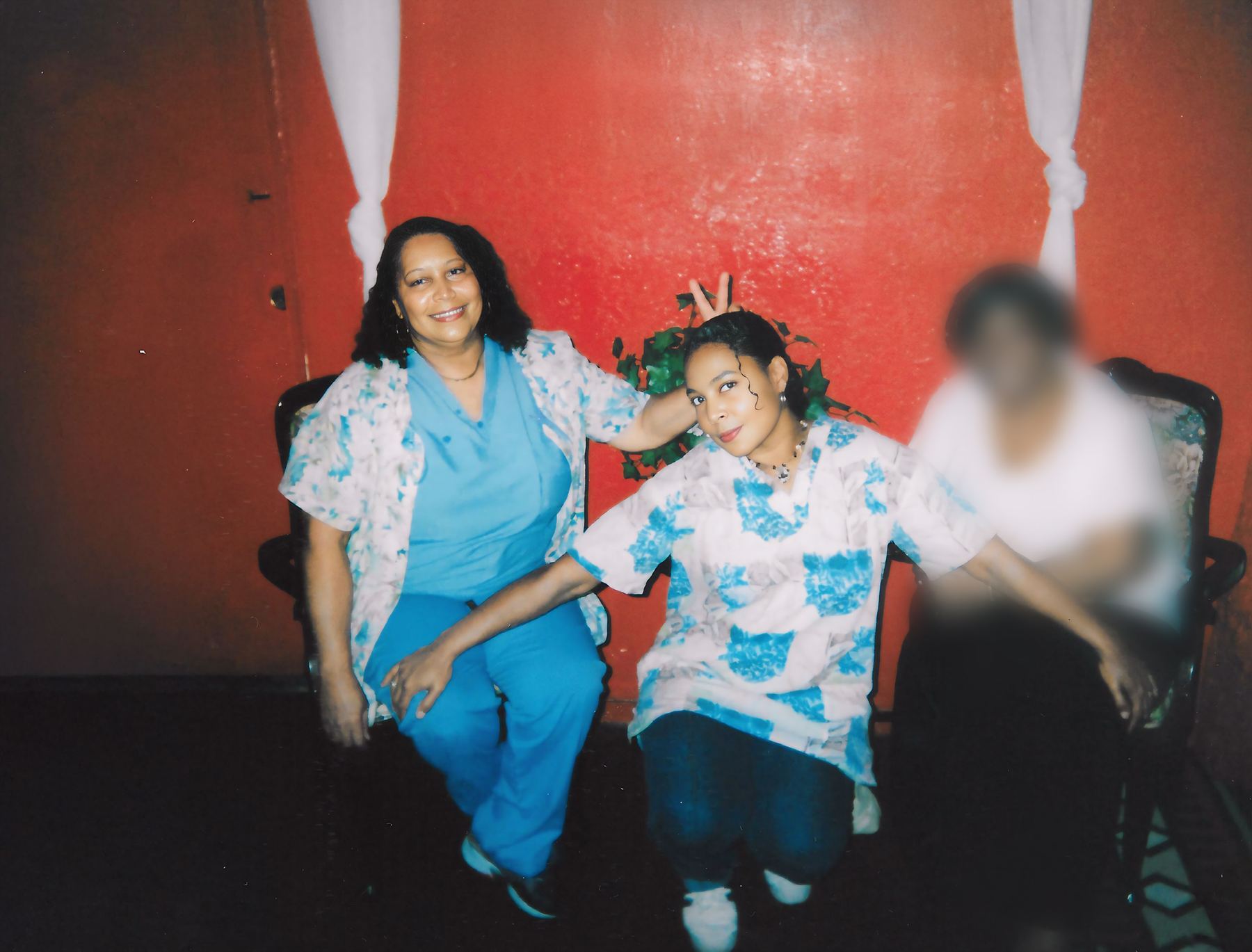 Lucretia Clark and her daughter Carla Taylor seated together against a red wall, Carla making bunny ears behind her mother's head.