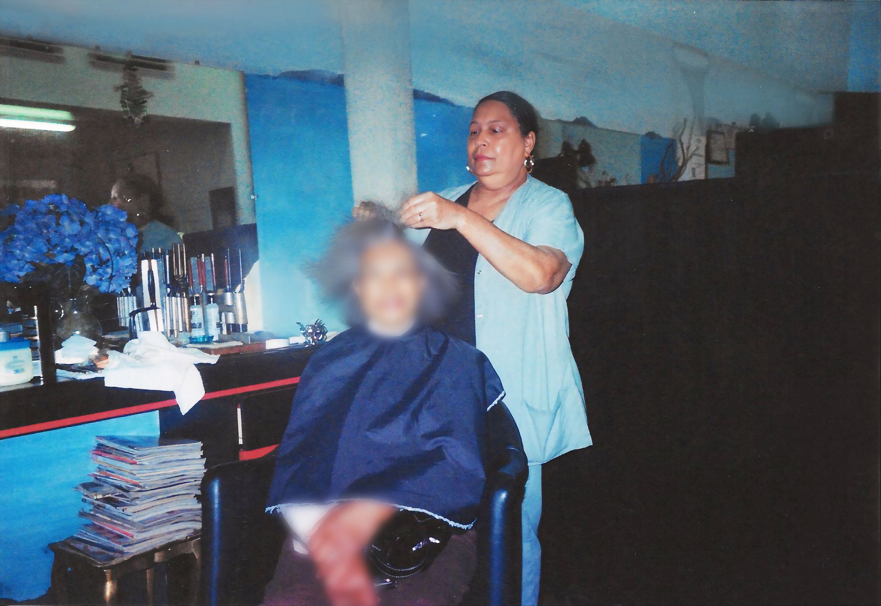Lucretia Clark styling a client's hair at her station, blue walls, a stack of magazines on the counter.