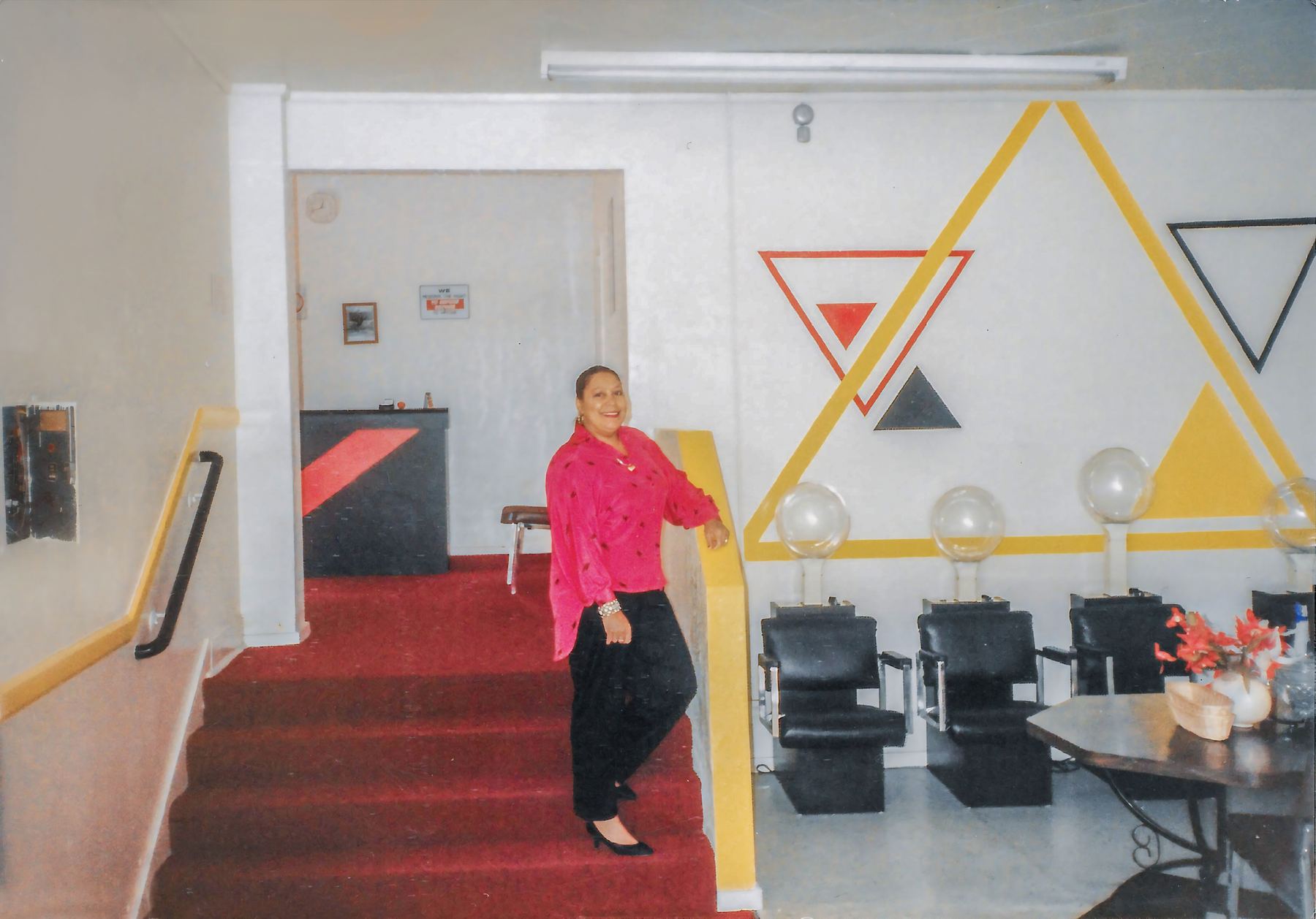 Lucretia Clark in a pink blouse, smiling on the red-carpeted steps of Renaissance Hair Salon, dryer chairs lined up beneath painted yellow triangles.